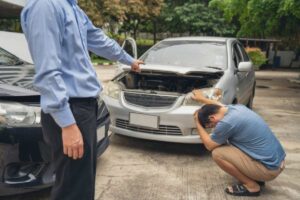 A sinistra una persona che punta il dito verso un auto con il cofano aperto e a destra un uomo accovacciato disperato